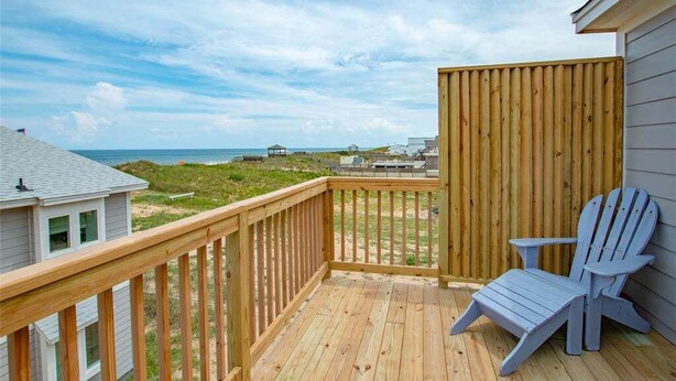 A wooden deck with a view of the ocean and dunes, a blue Adirondack chair, and a wooden fence.