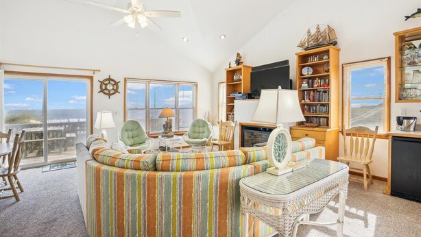 Un salon spacieux doté d’une grande fenêtre, d’un ventilateur de plafond, d’une bibliothèque et d’un coin repas offrant une vue sur la plage.