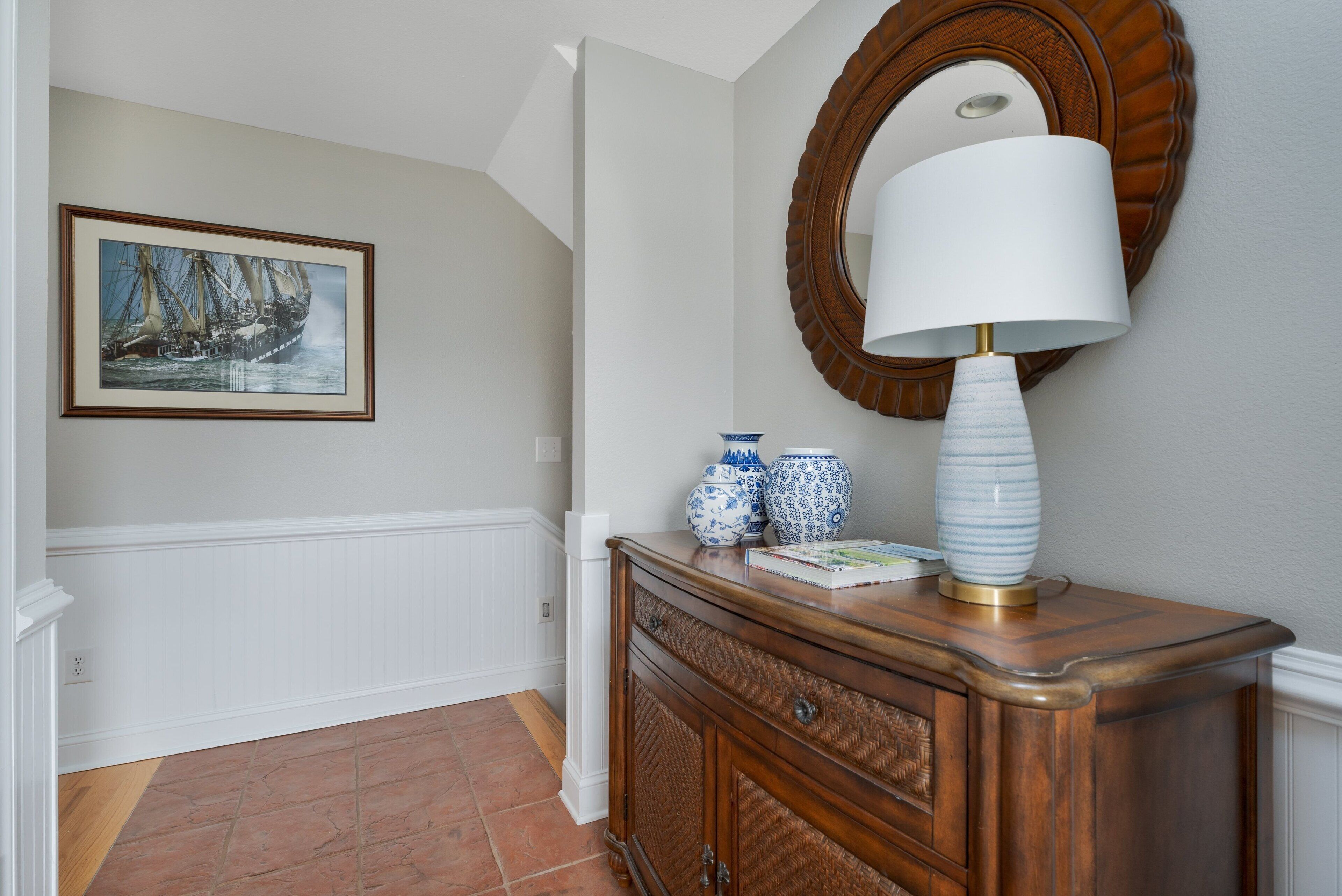 A hallway with a framed nautical painting, a wooden console table with a lamp, and decorative vases.