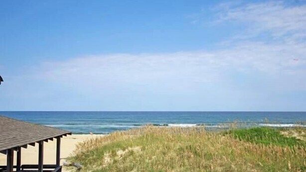 A beach view with a wooden structure, likely a beach hut, and a grassy dune area. - 5443- Gillam Cottage, Oceanfront | Nags Head | Sleeps 8 (Nags Head)