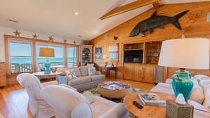 Un salon chaleureux avec un grand plafond en bois, un téléviseur et une vue sur l’océan grâce à de grandes fenêtres.