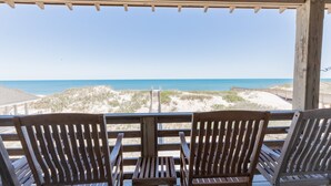 Une terrasse en bois avec deux chaises en bois, donnant sur une plage bordée de dunes de sable et l’océan.