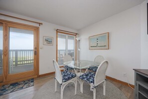 Een kamer met een glazen tafel en witte rieten stoelen, een houten deur en een lijst met een afbeelding aan de muur.