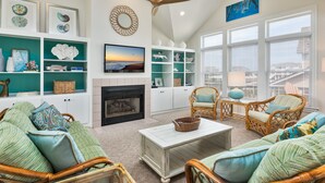 A spacious living room with a fireplace, wicker furniture, and a view of the city through large windows. - 5685- Seaside - Nags Head Coastal Home Near The Beach (Nags Head)