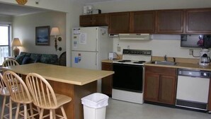 Cocina compacta con armarios de madera, estufa, refrigerador y lavavajillas. Zona de comedor con sillas y una mesa.