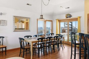 A dining room with a wooden table and chairs, a hanging light fixture, a mirror, and a window with blinds. - 5702- It's A Wonderful Life, Steps to Beach | Pool, Hot Tub & Game Room (Nags Head)