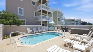 A pool area with lounge chairs, a hot tub, and a multi-story building in the background. - 5702- It's A Wonderful Life, Steps to Beach | Pool, Hot Tub & Game Room (Nags Head)