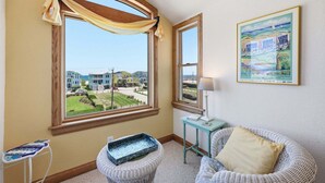 A room with a large bay window, a wicker chair, a small table, and a framed painting. - 5702- It's A Wonderful Life, Steps to Beach | Pool, Hot Tub & Game Room (Nags Head)