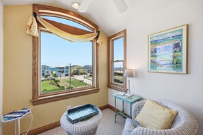 A room with a large bay window, a wicker chair, a small table, and a framed painting. - 5702- It's A Wonderful Life, Steps to Beach | Pool, Hot Tub & Game Room (Nags Head)