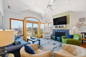 A living room with a fireplace, a television mounted above it, and a view of the city through large windows. - 5702- It's A Wonderful Life, Steps to Beach | Pool, Hot Tub & Game Room (Nags Head)