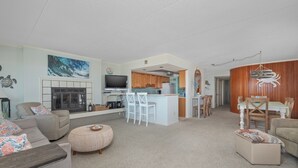 A spacious living room with a fireplace, a kitchen area with wooden cabinets, a dining table with chairs, and a small island with bar stools.