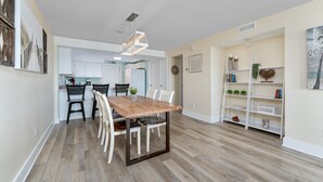 Un coin repas avec une table et des chaises en bois, une cuisine en arrière-plan et une bibliothèque agrémentée d’objets décoratifs.