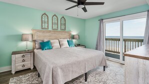 Une chambre à coucher comprenant un lit, des tables de chevet, un ventilateur de plafond, une fenêtre donnant sur la plage et des œuvres d’art décoratives au mur.