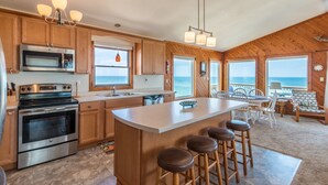 A kitchen with wooden cabinets, stainless steel appliances, a large island with stools, and a view of the ocean through large windows. - 5782-Whale of a View - Charming Oceanfront Nags Head Cottage (Nags Head)