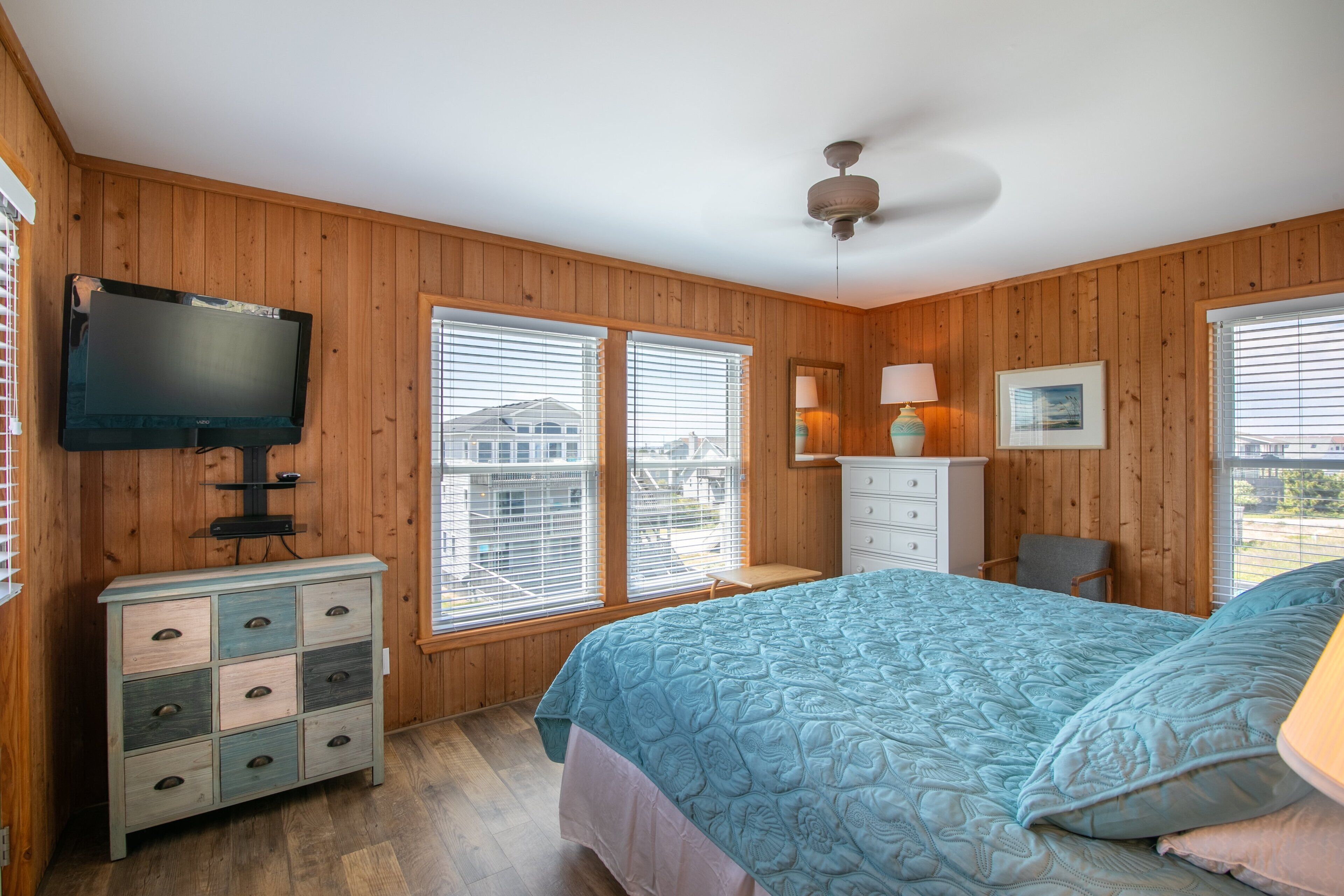 Un dormitorio con paredes de madera, una cama con colcha azul, un aparador, un televisor y dos ventanas con persianas.