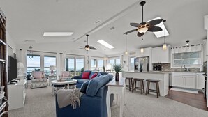 A spacious living room with a blue sofa, a dining area with a kitchen in the background, and large windows offering a view of the ocean. - 5786- After All - Oceanfront South Nags Head w/ Elevator (Nags Head)