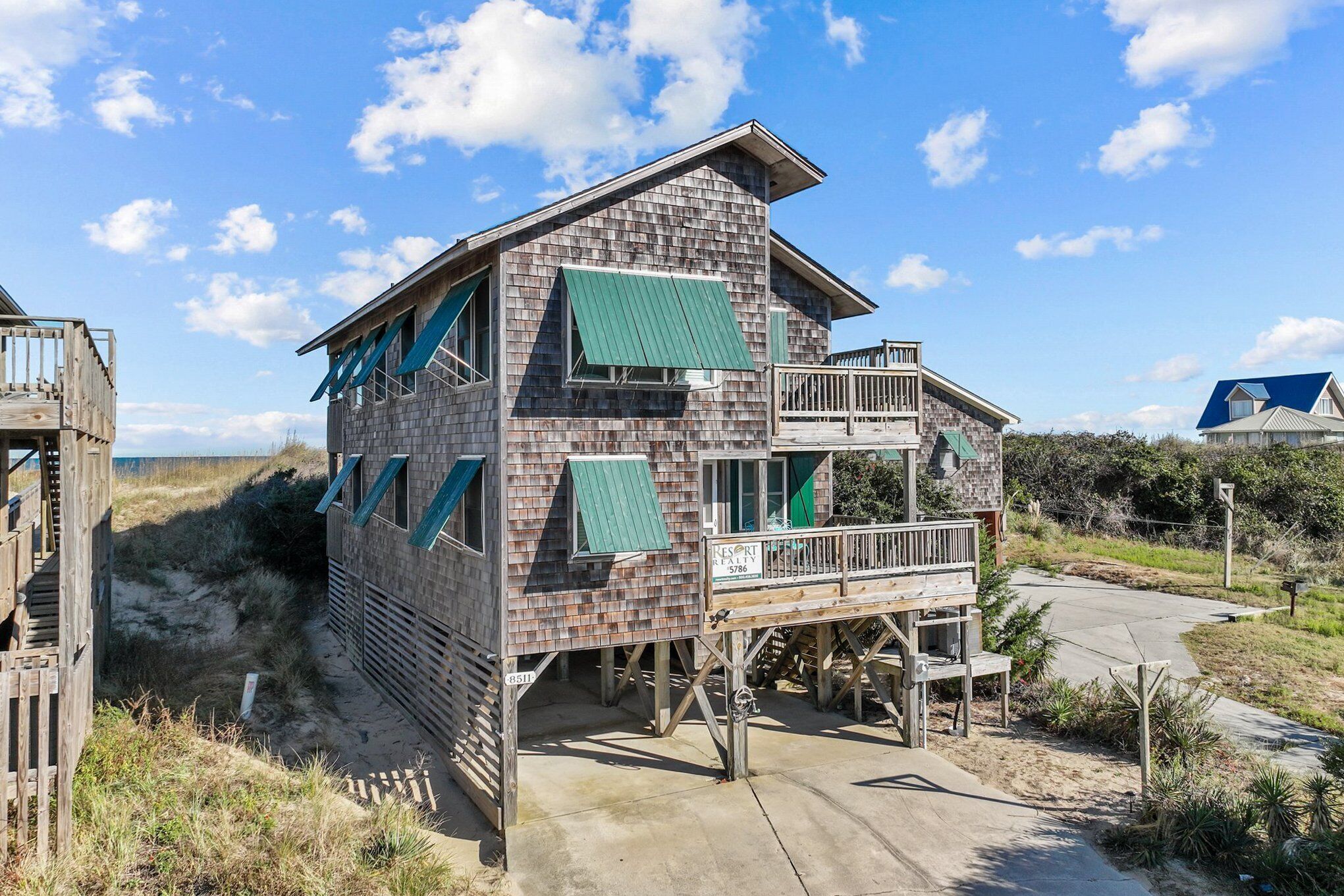 A two-story beach house with a balcony, a deck, and a sandy surrounding.