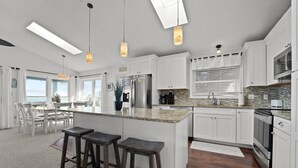 A modern kitchen with white cabinetry, a central island with stools, a stainless steel refrigerator, and a large window providing natural light. - 5786- After All - Oceanfront South Nags Head w/ Elevator (Nags Head)