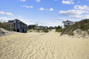 Un sentier de sable qui longe la plage et mène à des maisons situées en bord de mer, sous un ciel dégagé.