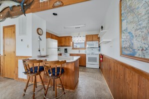 Une cuisine avec des meubles de rangement en bois, un réfrigérateur blanc, un four à micro-ondes et une cuisinière. On y trouve deux chaises en bois avec des coussins bleus.