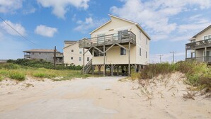 A two-story beach house with a balcony, a staircase leading to the upper level, and a sandy beach in the foreground. - 6504- Eagle's Nest - Quiet Ocean View Escape | Walk to Beach (Nags Head)