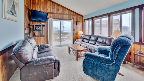 A living room with a brown leather sofa, a blue armchair, a wooden coffee table, and a television mounted on the wall. - 6504- Eagle's Nest - Quiet Ocean View Escape | Walk to Beach (Nags Head)
