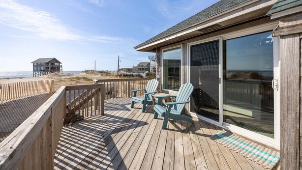 A wooden deck with two chairs and a table, overlooking a beach and a distant house. - 7026- Southwind - Rodanthe Cottage | 100 Yards to Beach (Rodanthe)