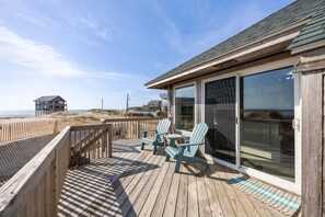 A wooden deck with two chairs and a table, overlooking a beach and a distant house. - 7026- Southwind, | Oceanside | Rodanthe | Sleeps 4 (Rodanthe)