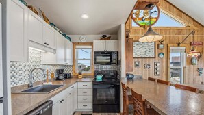 A compact kitchen with white cabinets, a black stove, and a wooden dining area. - 7026- Southwind - Rodanthe Cottage | 100 Yards to Beach (Rodanthe)