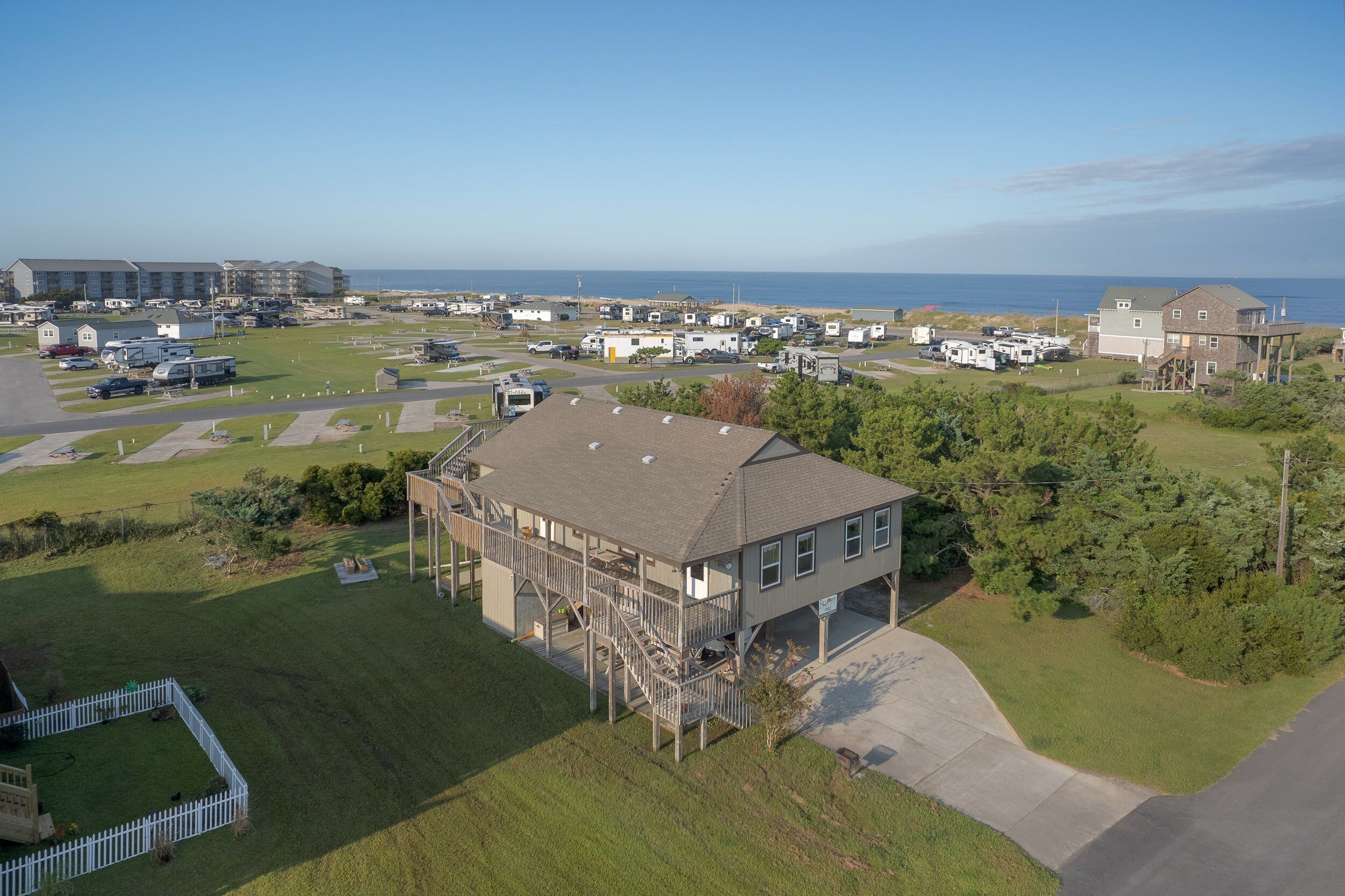 A residential area with multiple RVs parked on a grassy field, a large building with a balcony, and a view of the ocean.