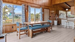 Un intérieur en bois comprenant une table à manger et des bancs, un coin cuisine et de grandes fenêtres avec des rideaux.