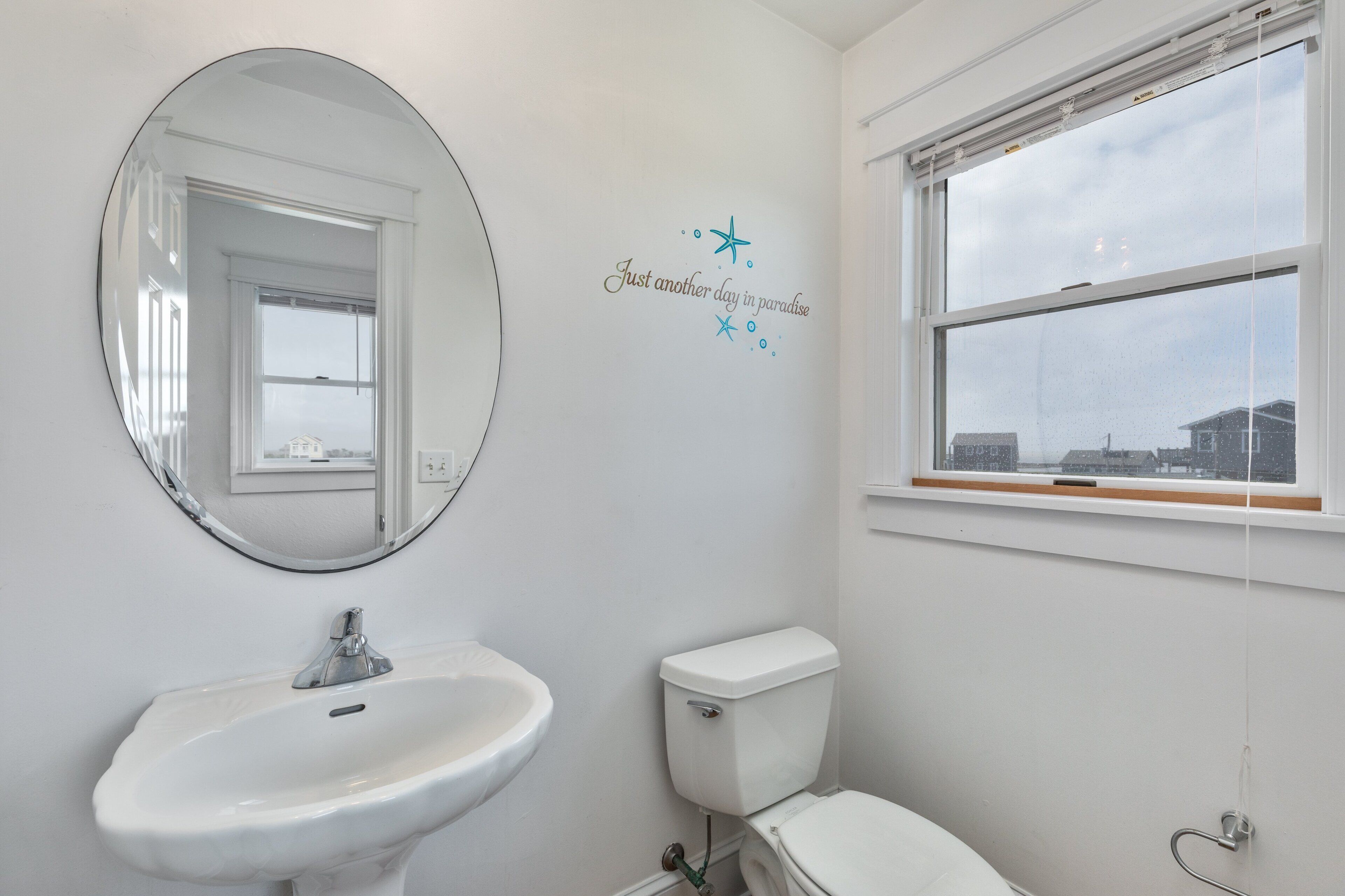 Un baño con lavamanos blanco, inodoro y un espejo grande. Se ve una ventana con vistas a edificios.