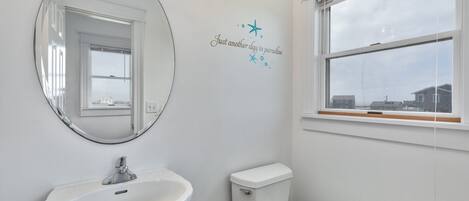 Un baño con lavamanos blanco, inodoro y un espejo grande. Se ve una ventana con vistas a edificios.