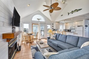 A spacious living area with a large blue sofa, a wooden coffee table, and a television mounted on the wall. There is a ceiling fan, a fireplace, and a view of the outdoors through a large window. - 7560-Sunset Cove - Relaxing Canalfront Retreat w/ Private Dock (Avon)