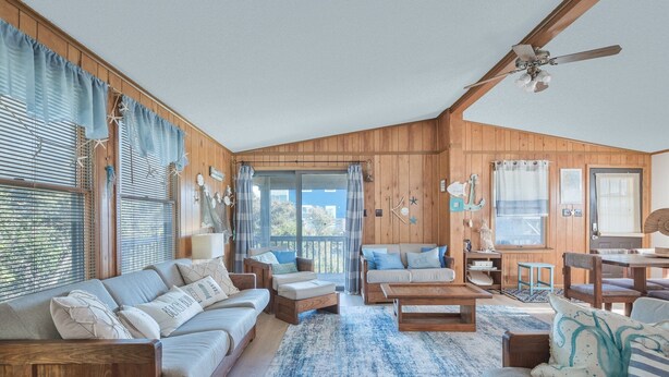 Une pièce confortable, aménagée comme un chalet, avec des murs en bois, un ventilateur de plafond, une grande fenêtre avec des rideaux, un canapé, une table basse et une petite table à manger.