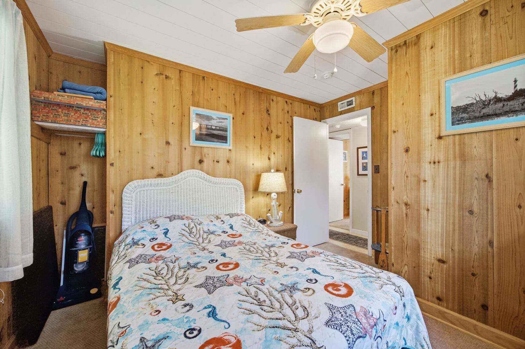 Una stanza con rivestimento in legno, un letto, una ventola a soffitto, una lampada e una porta che conduce a un'altra stanza.