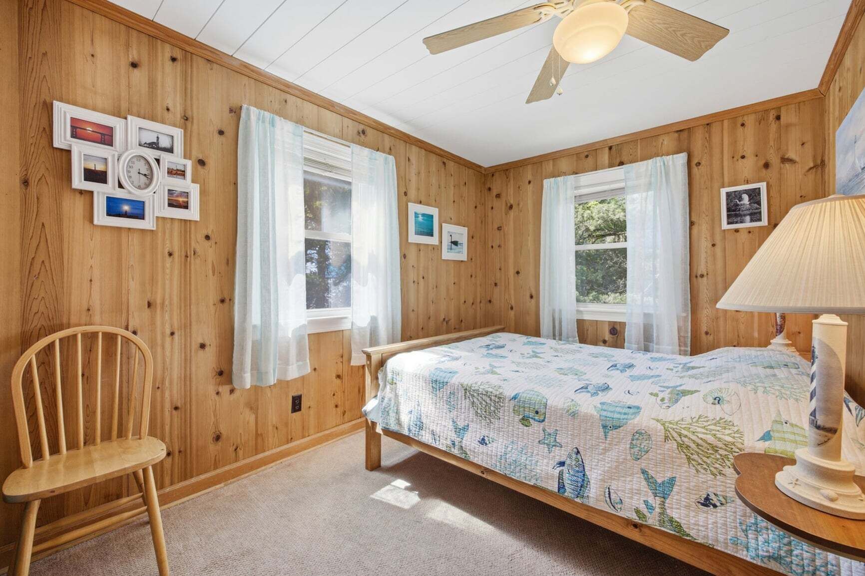 A wooden-paneled room with a bed, two windows with sheer curtains, a chair, and a lamp.