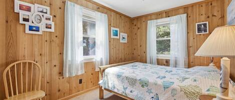 A wooden-paneled room with a bed, two windows with sheer curtains, a chair, and a lamp.