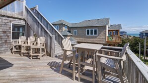 Una terraza de madera con sillas Adirondack y una mesa, con vista a la playa y otros edificios.