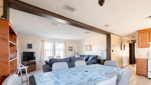 Ein modernes Wohnzimmer mit großem Fernseher, einer Couch, einem Esstisch mit Stühlen und einer Küchenzeile mit Holzschränken.