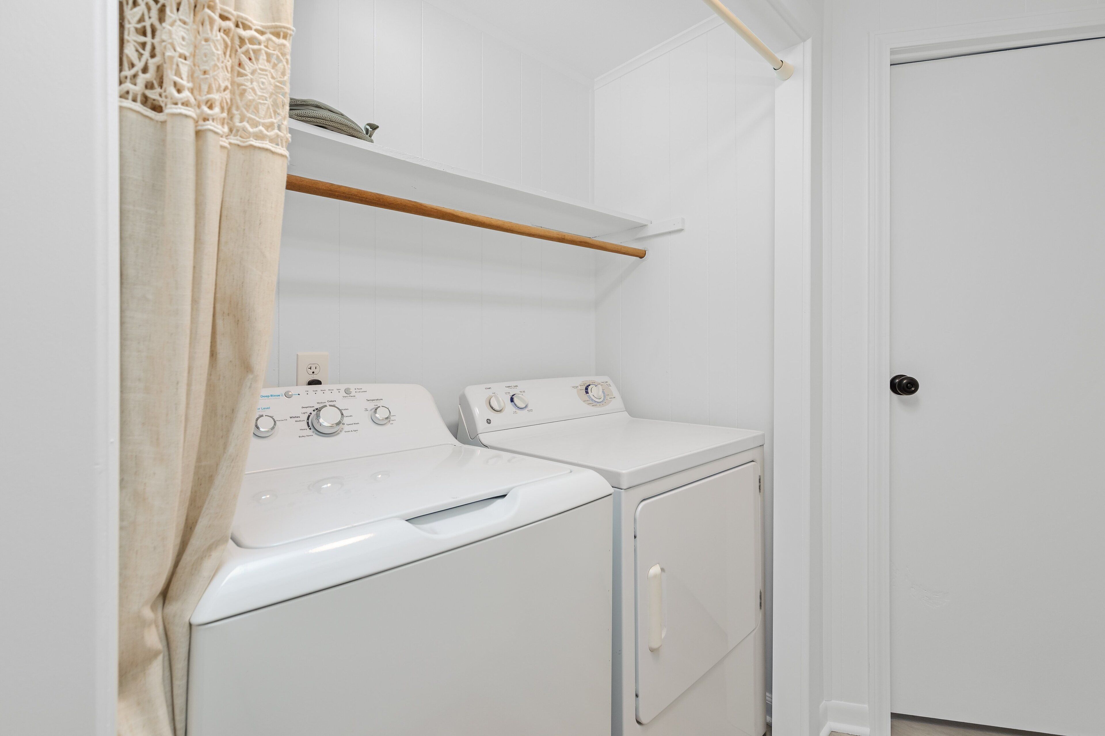 A laundry room with two white washing machines, a wooden shelf above, and a door to the right.