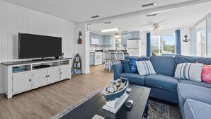 Una sala de estar con televisión, sofá y mesa de centro. Se observa una cocina al fondo con gabinetes blancos y un microondas.