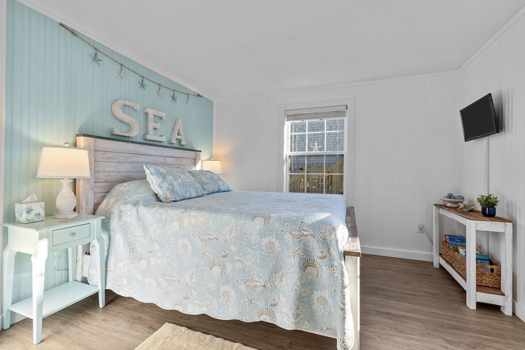 Un dormitorio con cama, mesita de noche, televisor montado en la pared y ventana decorada con una estrella de mar.