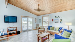 A spacious living room with a wooden ceiling, a flat-screen TV, a wooden coffee table, a striped sofa, and a large window with a view of the outside. - 7950- Ocean, | Coastal Hatteras Escape | Walk to Beach (Hatteras)