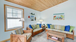 A cozy living room with a wooden ceiling, a large window with blinds, a striped sofa set, and a coffee table. - 7950- Ocean, | Coastal Hatteras Escape | Walk to Beach (Hatteras)
