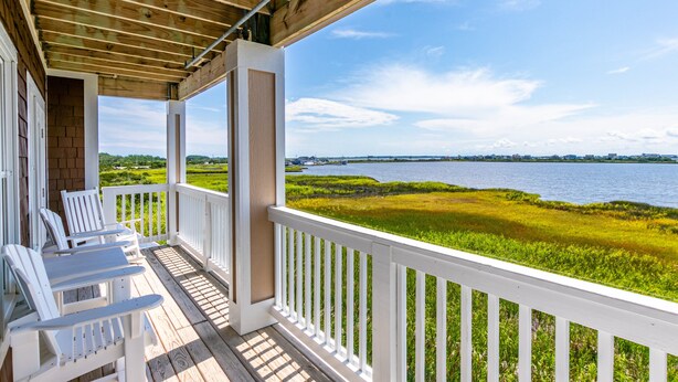 A wooden deck with white railings and chairs, overlooking a body of water and grassy areas.