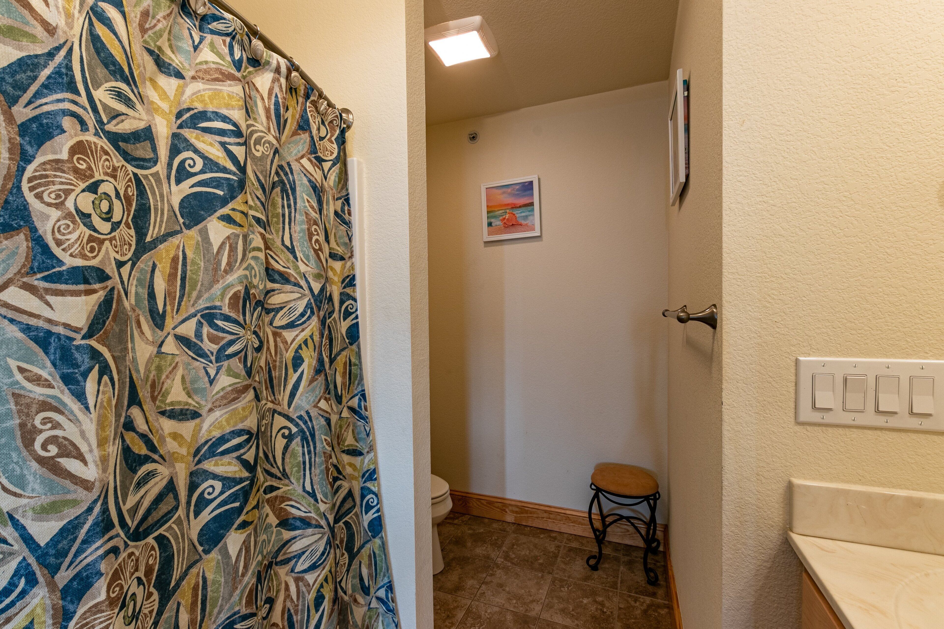 Une salle de bain avec un rideau de douche à motifs, des toilettes, un petit tabouret et un luminaire fixé au mur.