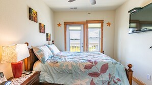 A bedroom with a bed, a nightstand with a lamp, a ceiling fan, and a TV mounted on the wall.