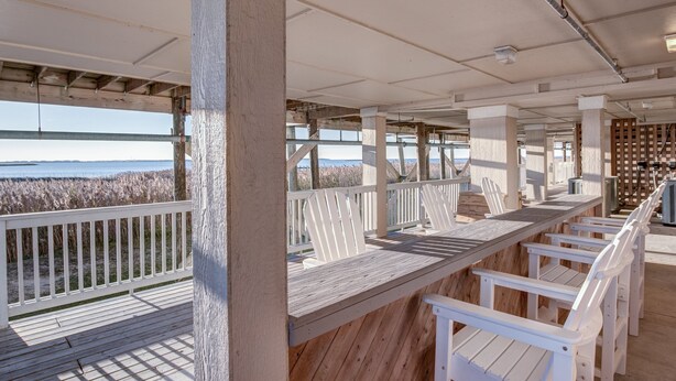 A covered outdoor seating area with wooden tables and white chairs, overlooking a beach with tall grasses. - C213-Sound and Sea 213, | Soundfront | Sugar Creek | Nags Head | Sleeps 6 (Nags Head)