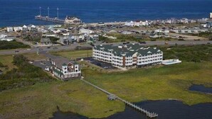 An aerial view of a coastal town with a prominent multi-story hotel, a pier extending into the water, and a mix of residential buildings. - C213-Sound and Sea 213, | Soundfront | Sugar Creek | Nags Head | Sleeps 6 (Nags Head)